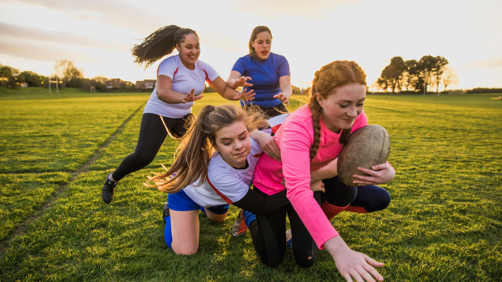 3 teen girls tackling another girl with a rugby ball